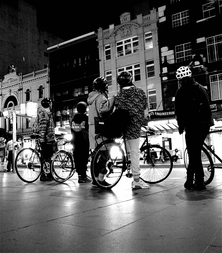 Black and white photograph, of 3 cyclists stopped on a city street, looking up at a projections on city buildings in Melbourne.