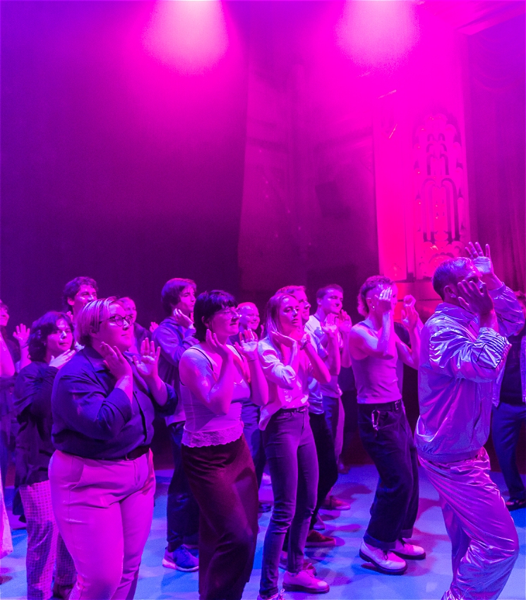 A group of people led by Joel Bray dance on a dancefloor. All the people are lit by a purple light.