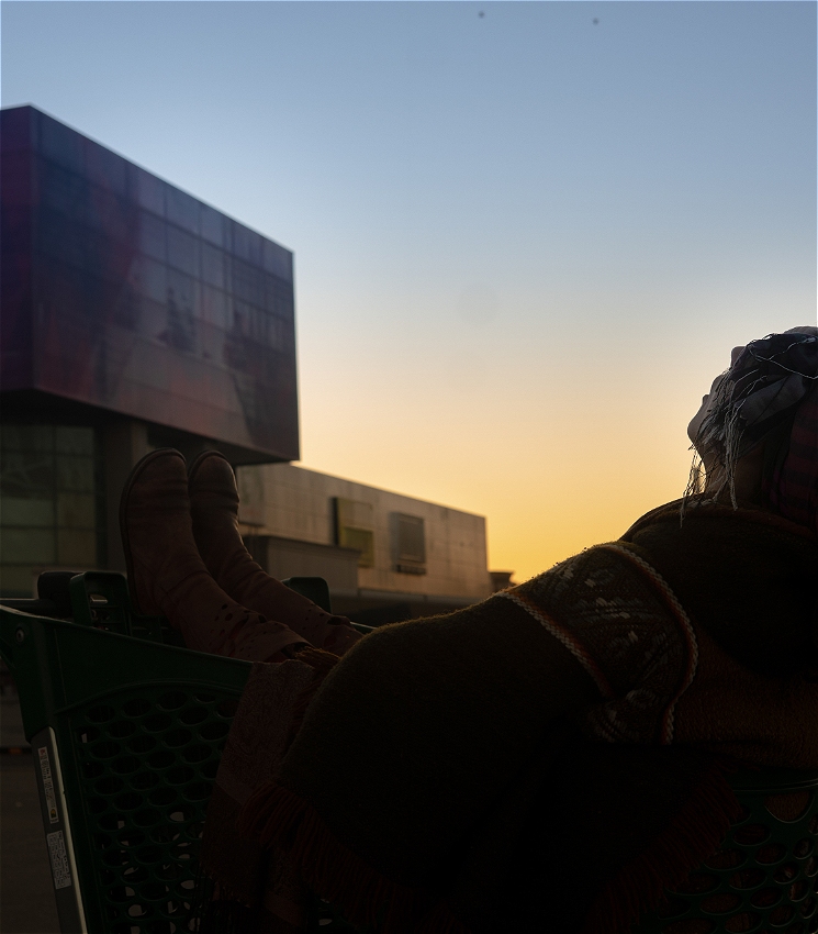 Sunrise outside in a shopping center carpark. In background the orange sun peaks from the horizon, emerging behind a shopping center complex. In the foreground is the silhouette of a woman siting inside a shopping cart, exhaling into the cold blue sky. 
