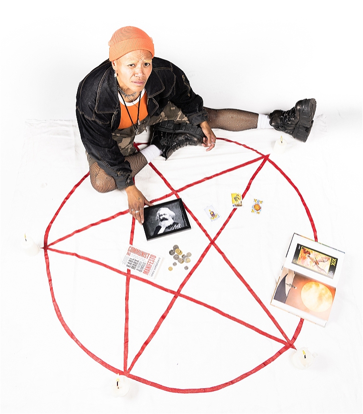 A dark skinned femme person sitting on a white background. They are wearing a long brown skirt, an oversized black sport jersey, a pink knitted beanie, and black chunky boots. They have bleached eyebrows, and are looking sceptically into the camera. There is a red pentacle painted on the ground. Around them are various witchy items such as candles, books, and crystals. In the centre of the pentacle is a pile of coins, a copy of 'The Communist Manifesto', and a framed photo of Karl Marx.