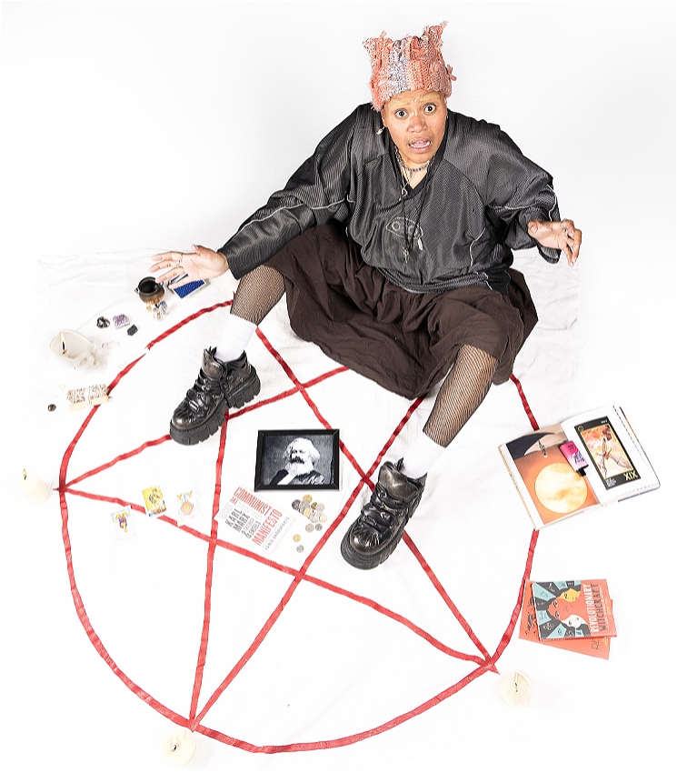 A dark skinned femme person sitting on a white background. They are wearing a long brown skirt, an oversized black sport jersey, a pink knitted beanie, and black chunky boots. They have bleached eyebrows, and are looking shocked at the camera. There is a red pentacle painted on the ground. Around them are various witchy items such as candles, books, and crystals. In the centre of the pentacle is a pile of coins, a copy of 'The Communist Manifesto', and a framed photo of Karl Marx.