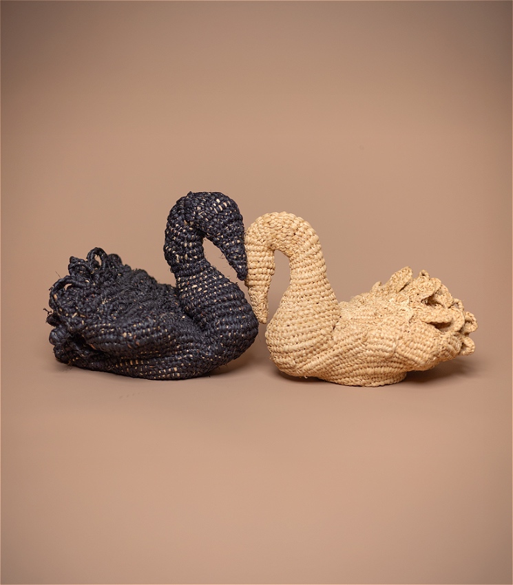 A close up photograph of two swans, one black the other beige, made out of woven natural material, in front of a tan paper backdrop. The swans are facing each other, kissing.
