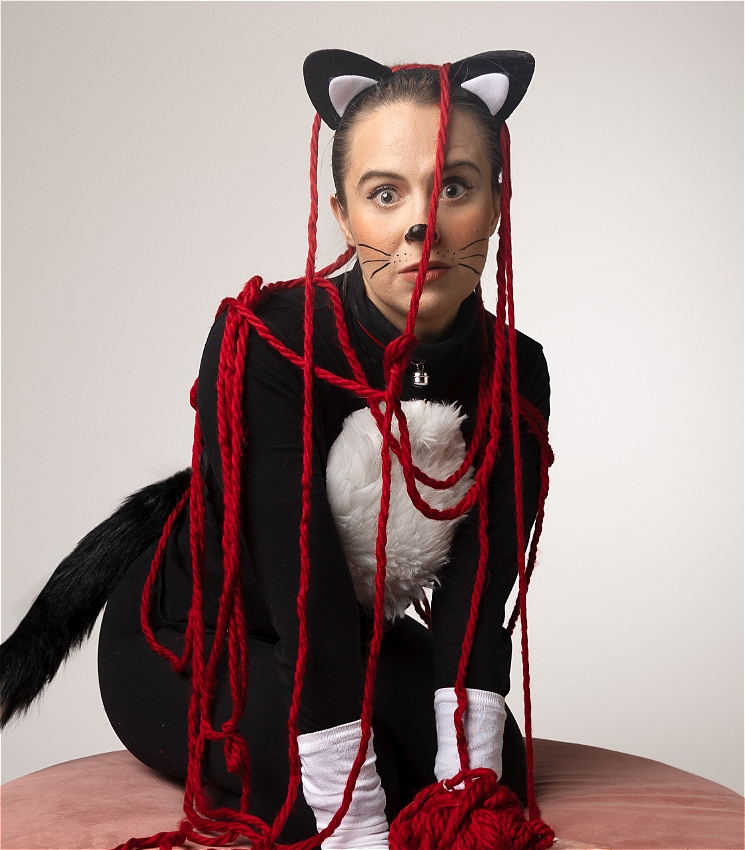 A still image of a woman wearing a cat costume covered by strands of red yarn. She looks at the viewer wide eyed while her hands (as paws) rest on a pink cat bed with a red ball of yarn.