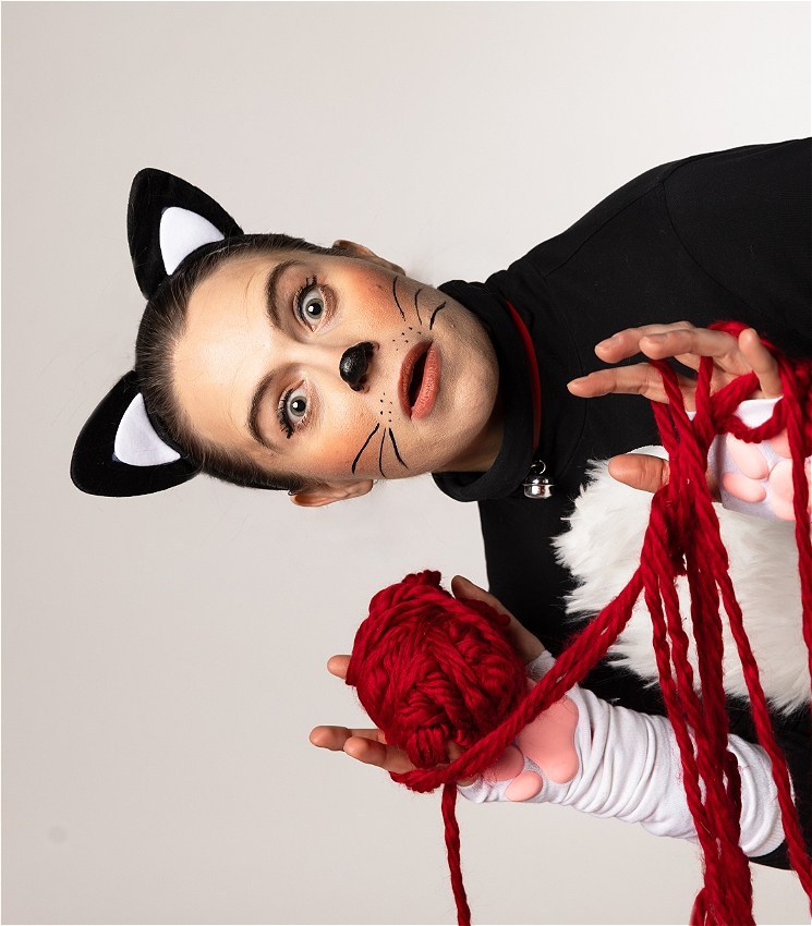 A sideways image of a woman wearing a cat costume and holding a ball of red yarn, looking at the camera with her paw-shaped hands playing with the yarn.