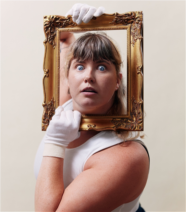  A girl with long hair and blunt bangs holds an ornate gold picture frame around her face. She is wearing white gloves and a white sleeveless top. Her eyes are wide open, and she has a surprised expression.
