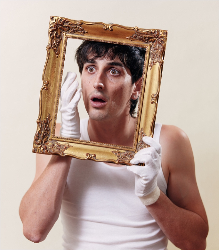 A boy with short dark hair holds an ornate gold picture frame around his face. He is wearing white gloves and a white sleeveless top. His mouth is slightly open, and he has a surprised expression. 