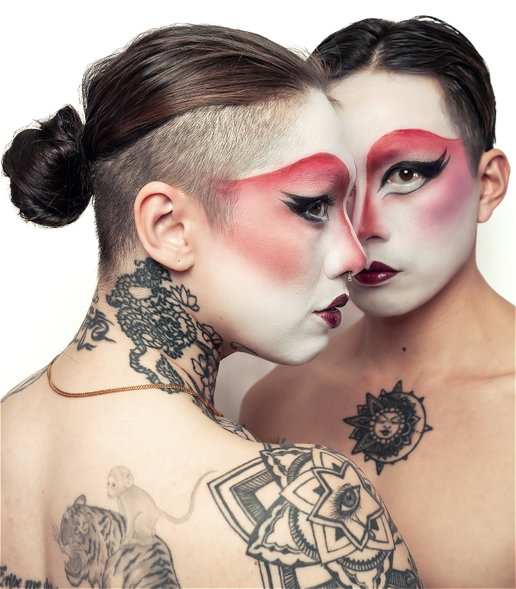 Charlie and Dyan, two genderfluid performers of Chinese descent stand face to face, their foreheads and noses almost touching. Both wear striking drag makeup inspired by Chinese opera. White base, bold red eyeshadow that sweeps across the brows, dramatic winged eyeliner, and dark lips. Their hair is slicked back, one in a low bun with shaved sides. Their bare shoulders and backs are visible, decorated with large black-and-grey tattoos — floral mandalas, an eye, a tiger with a monkey on Charlie, and a sun design on Dyan's chest. The overall effect is intense and intimate, blending softness with power.