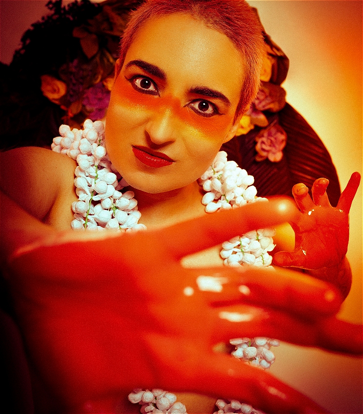 A performer, eyes-wide, stares ahead. Their hands covered in red paint, with floral wreaths around their neck and warm coloured flowers in the background.