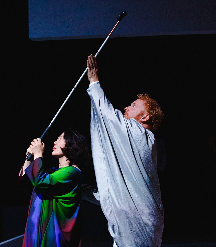 Two performers in close proximity. One performer is dressed colourfully and holds a cane above their head, the other performer reaches to touch it as though worshipping.