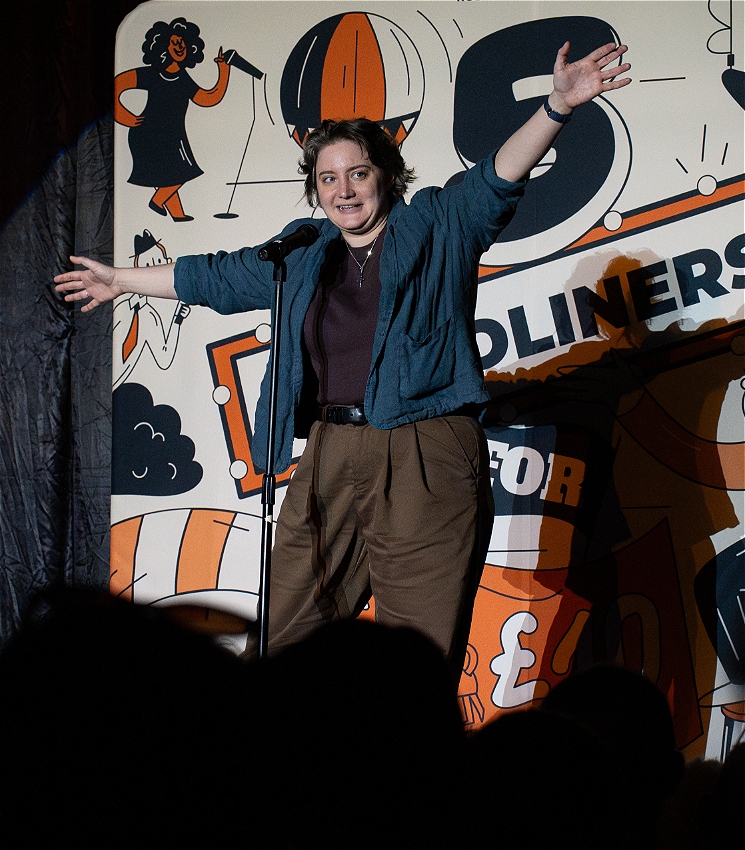 A white fem person is performing on stage with their arms stretched out wide , speaking into mic and eyes pointing to the audience. They are wearing brown trousers, a blue over shirt atop a plain black t-shirt. Their backdrop is a large 5 headliners for $25 image with several graphic images and text in a playful font.