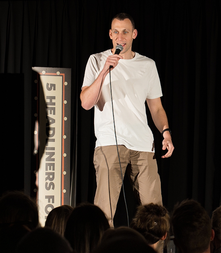 A white male is performing on stage, facing and engaged with audience, holding and speaking into a mic. They are wearing brown trousers, and a white t shirt.