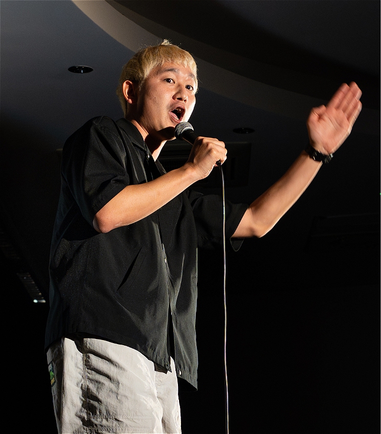 A photo of Takashi, a japanese comedian and performer, seen performing on stage, engaged with audience, and speaking into a mic. They are wearing great trousers, a black t shirt and have dyed golden hair.