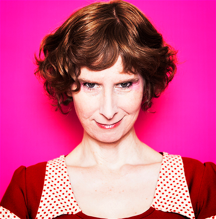 Telia Nevile, a woman with short, curly red hair, smiles menacingly at the camera, her head tipped down. She is shown from the shoulders up, wearing a red dress with a polka dot collar. The background is hot pink.