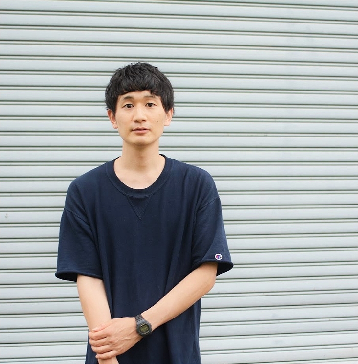 A person in blue tshirt and short hair stands in front of a white wall that looks like a roller door.