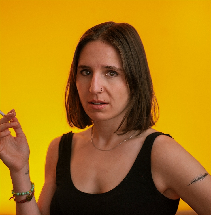 A person with shoulder length brown hair holds a cigarette and looks at the camera with a direct stare. There is a yellow background and they are wearing a black singlet.