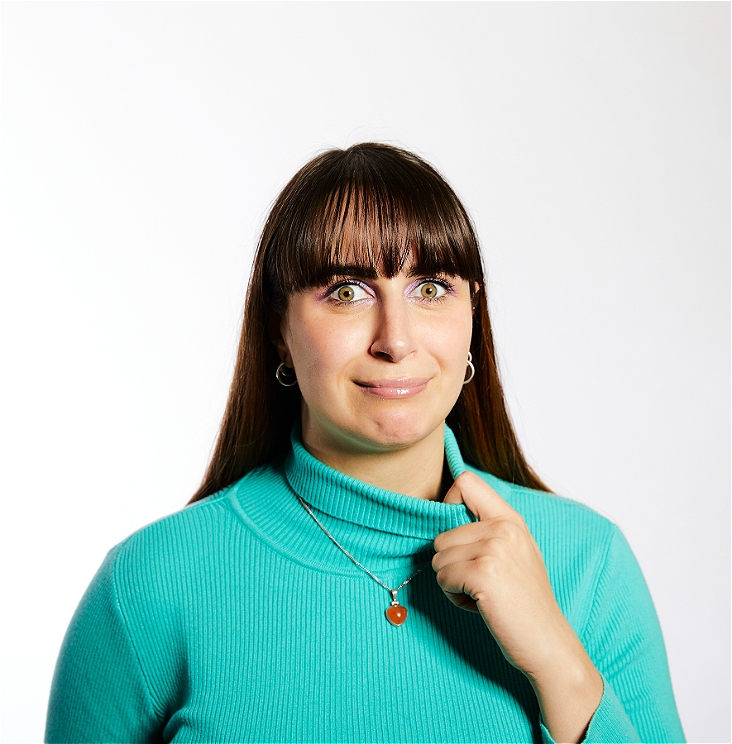 Ashley stands in front of a white background, wearing a bright teal turtleneck - she pulls at it's neck while staring into the camera with a concerned look in her eye.