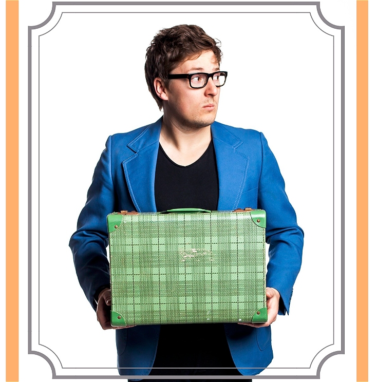 Angus, wearing a blue blazer, is looking off camera quizzically while holding a green suitcase in front of his chest.