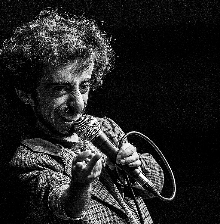 A black and white picture of Imaan mid performance with a cheeky grin. He is holding a microphone in one hand, and gesturing with his other hand.
