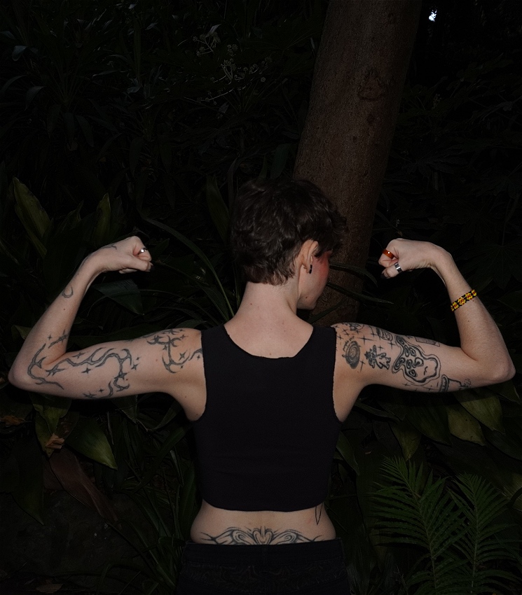 A trans masc person has their back to the camera and flexes with their arms in a pose. They have lots of tattoos, and are wearing a black binder and a bracelet with the Aboriginal flag colours. They are standing in the rainforest.