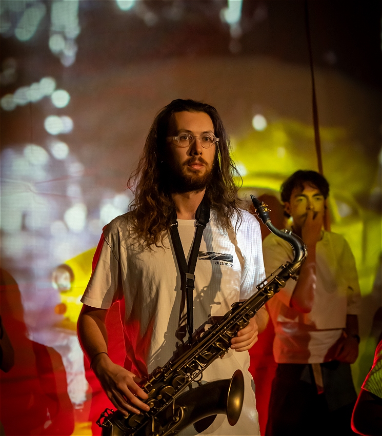 A waist-up photo of a young white man on stage (Toby Leman) with long brown hair and glasses. Toby is wearing a white t-shirt and holding a saxophone; his mouth remains closed while he stares intensely into the distance. The stage is lit in red and yellow, which creates orange-red shadows of Toby, while his saxophone glows gold. There is another masculine white person, standing in the background who is covering their mouth with their hand.