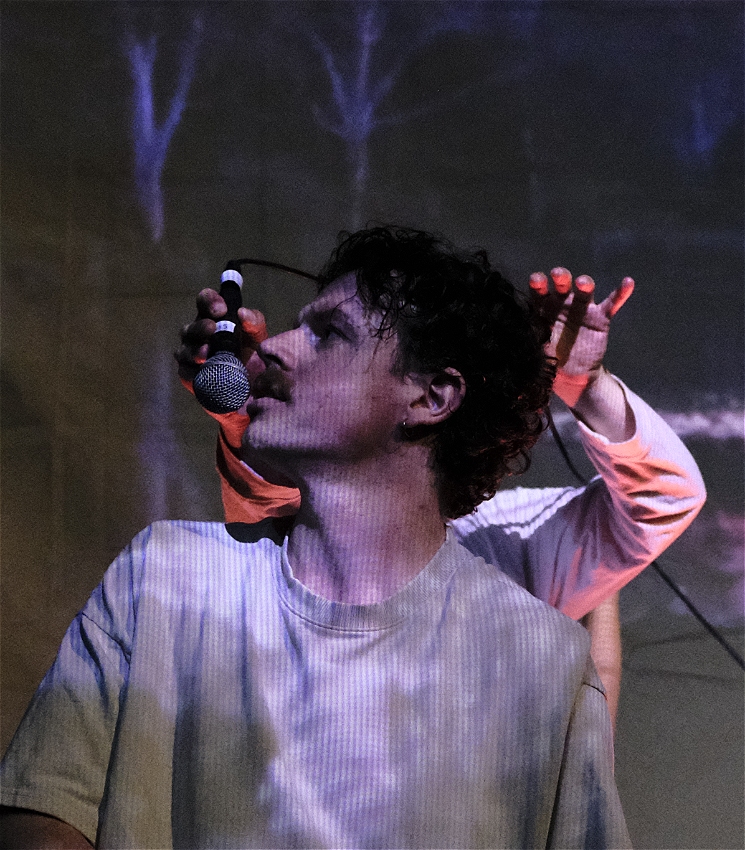 A man in his twenties (Ben Ashby) with short brown hair and a moustache is whispering into a microphone that is being dangled in front of his mouth by someone standing behind him. All we can see of the person behind him are their hands emerging from either side of his head.