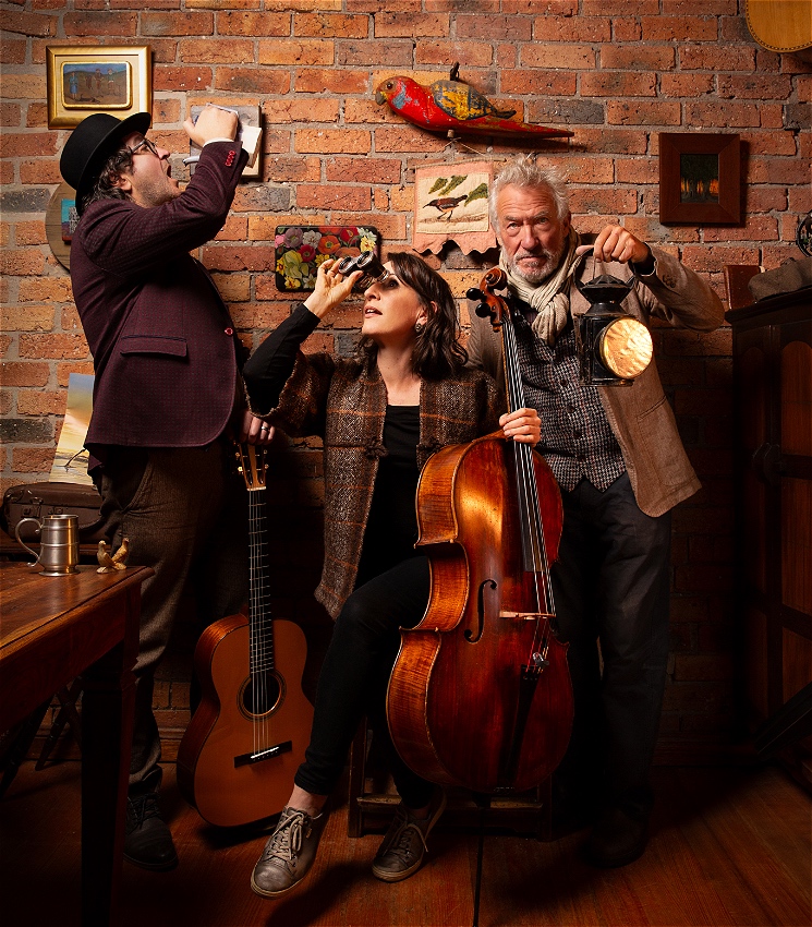 Three people are in a small room in front of a brick wall. A middle aged man on the left wearing a bowler hat and a jacket holds a guitar and appears to be drinking from a jug. In the middle is a younger woman in a knitted jacket holding a cello and looking into the distance through a pair of binoculars. On the right is a stern looking older man with grey hair and a beard wearing a waistcoat, scarf and jacket. He is holding a lantern. On the brick wall are various pictures of flowers and birds, and there is also a large wooden rosella.