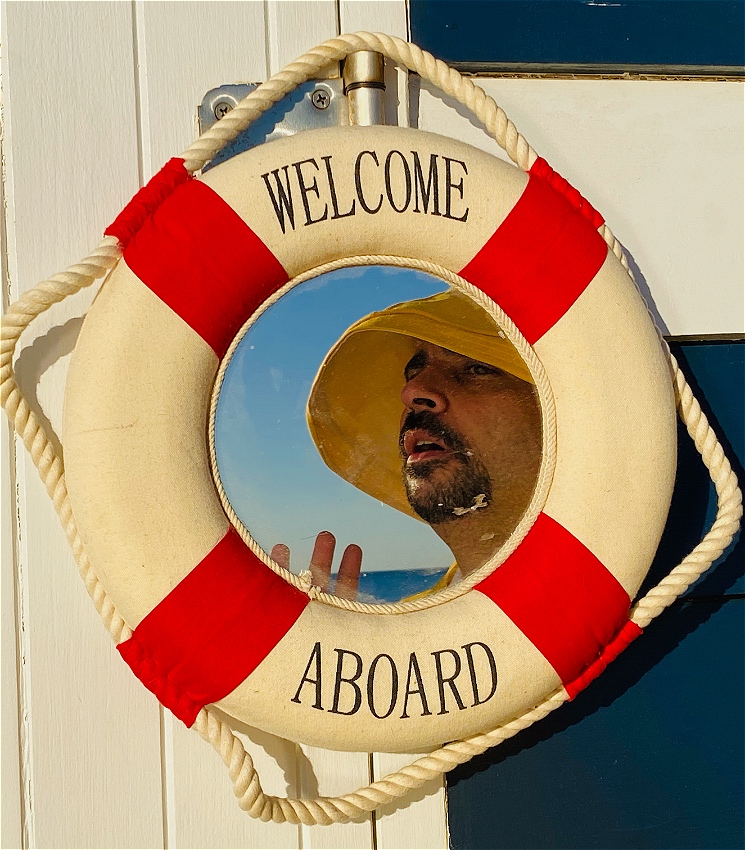 The reflection of a red and white striped lifesaver buoy-shaped mirror shows Jeremy Moses looking longingly with his hand reaching out. He is wearing a yellow raincoat and hat. A blue sky and sea are visible. The mirror is dirty, and says "WELCOME ABOARD".