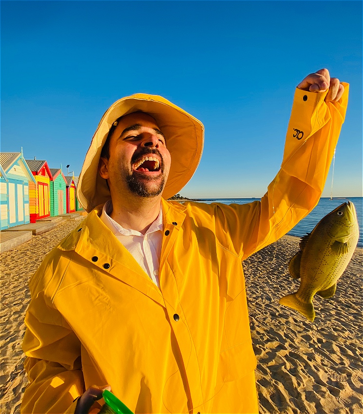 Jeremy Moses stands on a beach in front of a row of colourful wooden bathing boxes at sunset. He is wearing a yellow raincoat and hat, and is holding a plastic fish on a fishing line. He is open-mouth smiling.