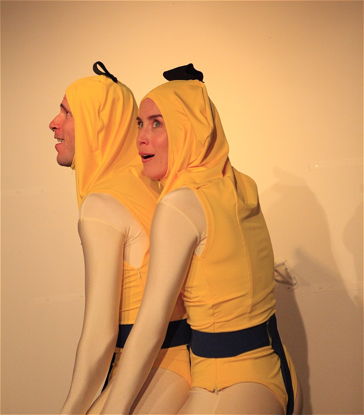 Two actors, dressed as bananas, bunching together in an art gallery with expressions of delight.