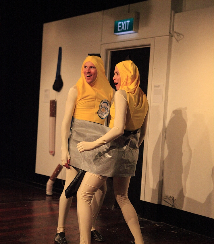 Two actors dressed as bananas are stuck together with duct tape, standing in an art gallery and looking at the audience with expressions of excitement.