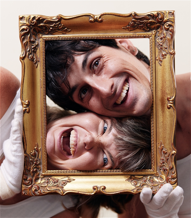 Two mimes hold an ornate golden picture frame. They are smiling / grimacing enthusiastically. An AMAB and an AFAB persons face are inside the frame and the look outwards off frame.
