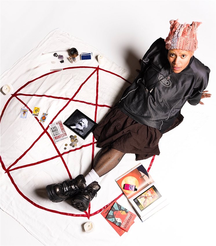 A dark skinned femme person sitting on a white background. They are wearing a long brown skirt, an oversized black sport jersey, a pink knitted beanie, and black chunky boots. They have bleached eyebrows, and are looking inquisitively into the camera. There is a red pentacle painted on the ground. Around them are various witchy items such as candles, books, and crystals. In the centre of the pentacle is a pile of coins, a copy of 'The Communist Manifesto', and a framed photo of Karl Marx.
