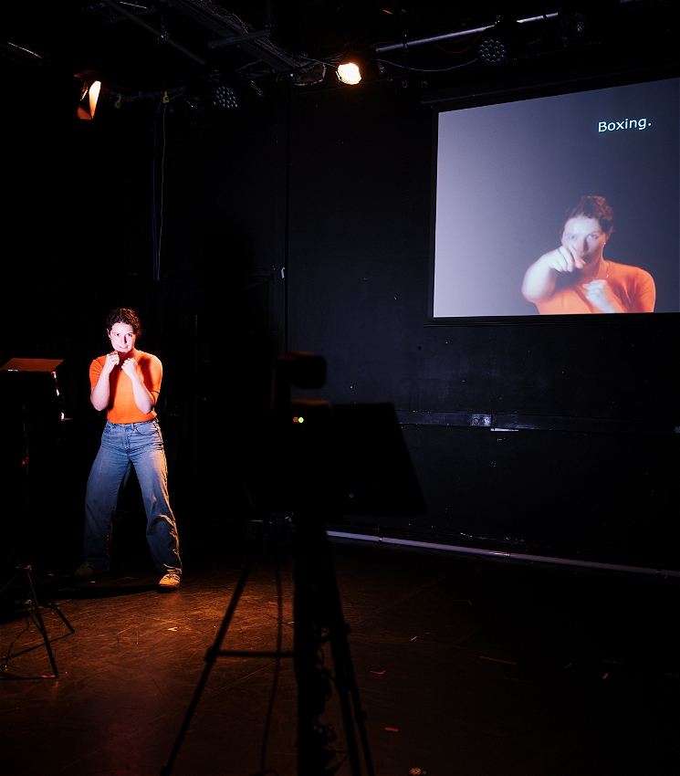 A person wearing jeans and an orange sweater stands at the back of the theatre, poised in a boxing stance. They face a camera which projects an image of their face onto a large screen above. The screen displays text that reads: boxing.