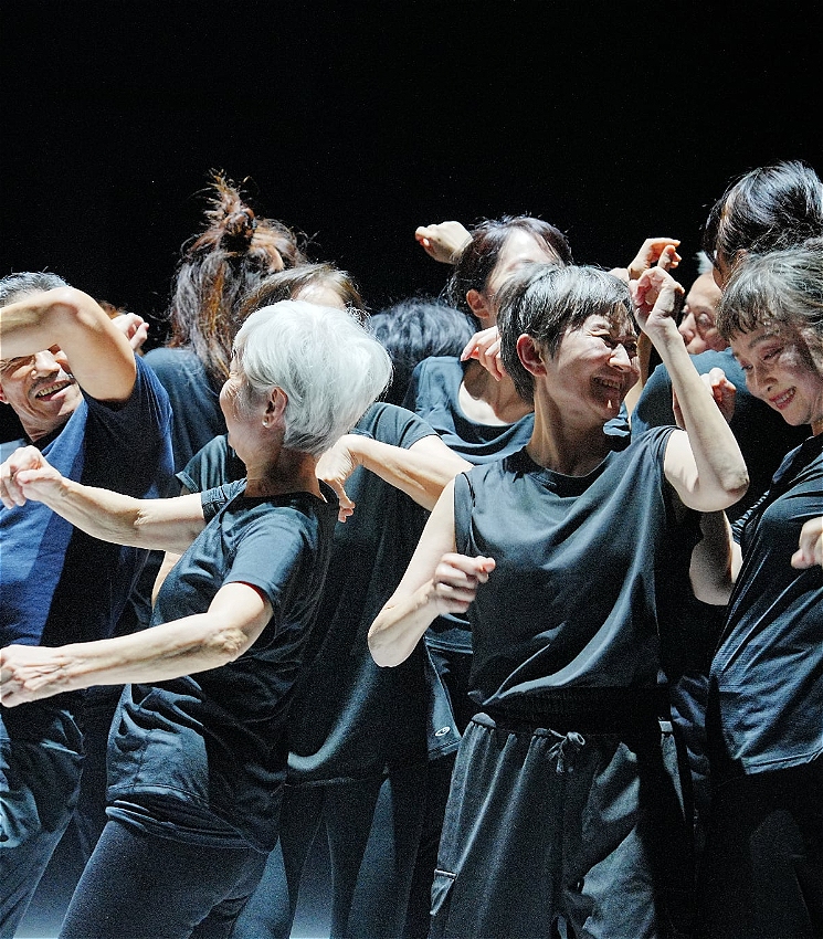 A huddle of dancers look to each other, smiling. They are all older performers, wearing all black, with their arms moving in interesting shapes.