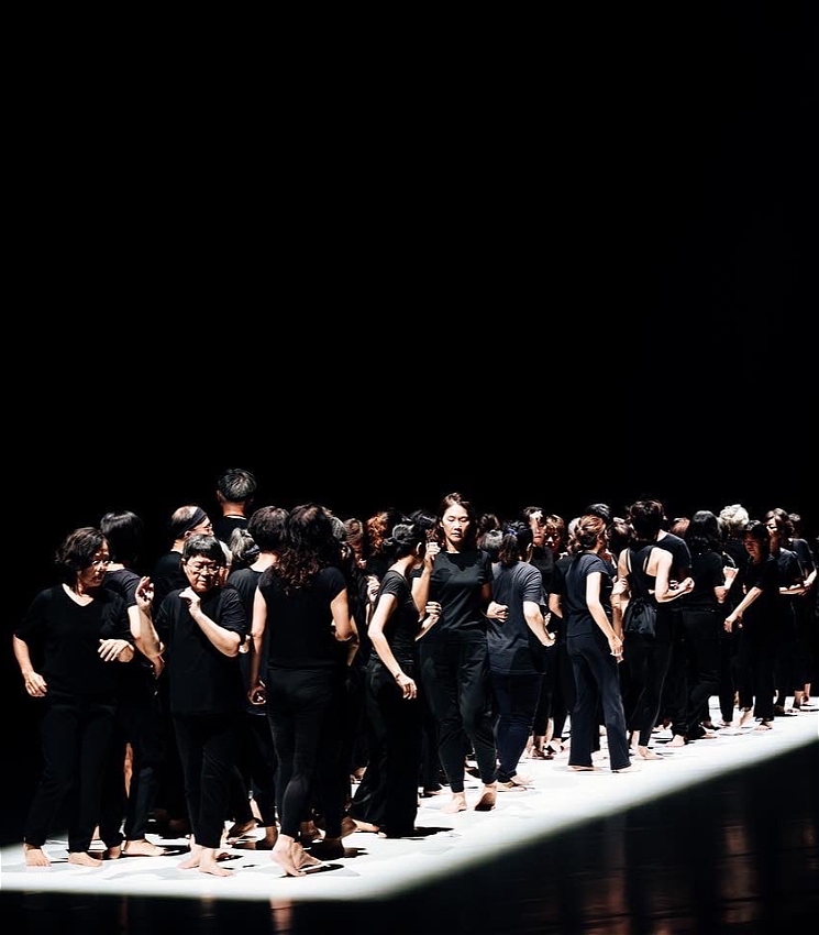 Against a stark black backdrop, a rectangle of light comes down from above. Within that rectangle are a mass of performers, all moving and walking past each other with stern or neutral looks on their faces. They are all wearing black, with their arms and feet exposed against the white floor.