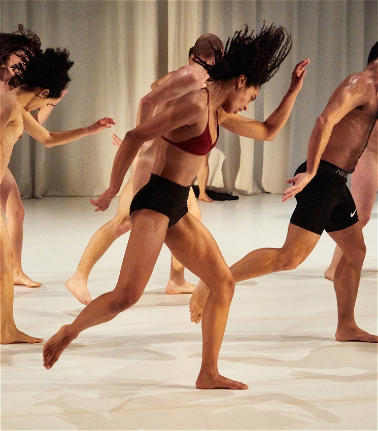 A group of mostly naked performers move in a synchronised motion with one arm in front of them and one arm behind them. They are against a stark white background.