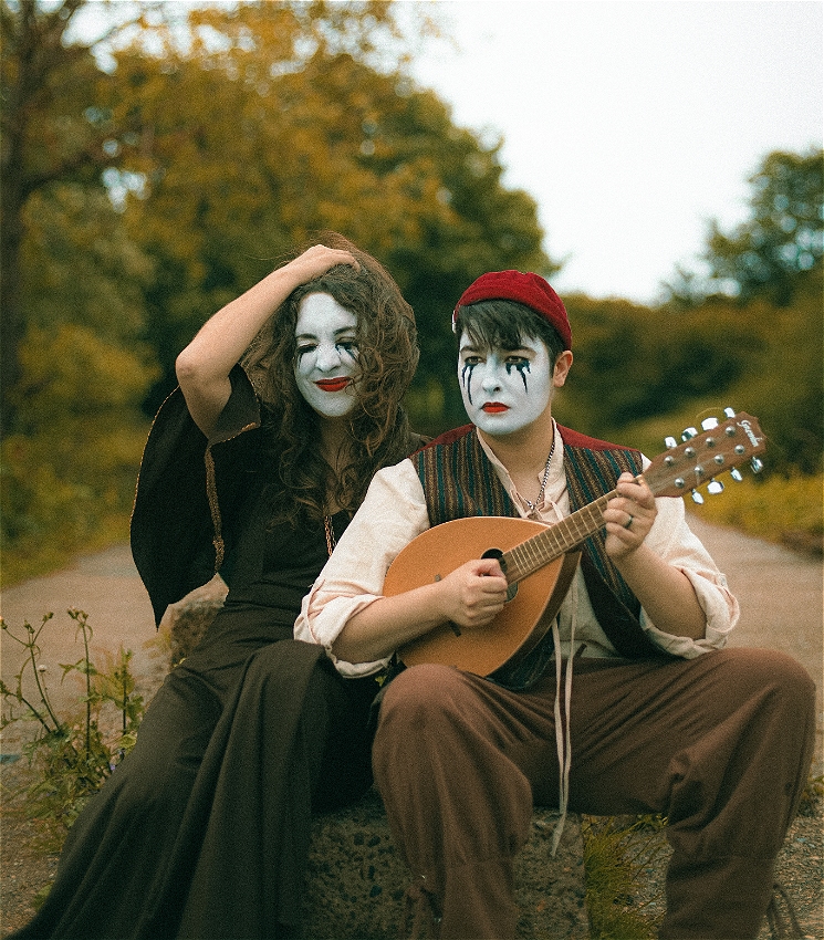 Two women sit side by side on a grey stone on a path. Their faces are painted white with black streaked mascara. The woman on the left has long curly hair and is wearing a brown dress, her hand is in her hair. The woman on the right has short hair, a red cap and is holding a mandolin. She is wearing trousers, a shirt and a waistcoat. There are trees in the background.