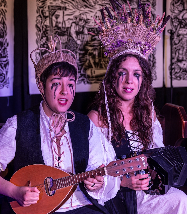 Two women with streaked black mascara are dressed in black and and are wearing headdresses made of barley. One woman plays a concertina and the other woman plays a mandolin. There are tapestries hung on the wall in the background.