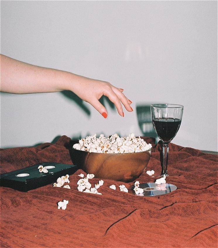 A flash photo taken with a film camera.
In the middle of the shot is a bowl of popcorn and a hand reaching down to grab some of the popcorn. The bowl is on a table with a red tablecloth.
On the left of the bowl is a black VHS, and on the right of the bowl is a retro crystal wine glass with red wine and a blue reflective DVD.