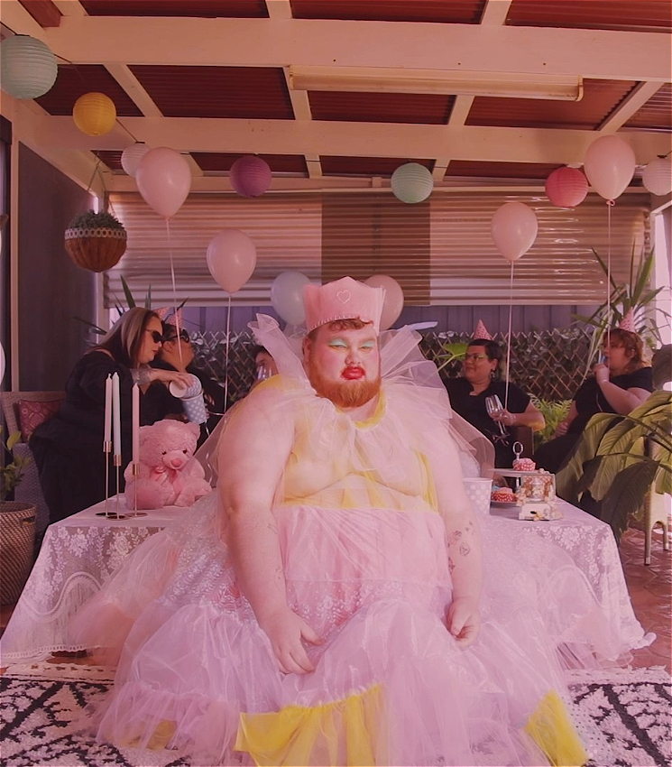 A person in a pink tulle dress, crown, and bold makeup sits with a dour expression. In the background other people are having a party with balloons.