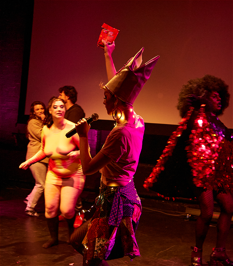A person wearing a pope hat with their back to the camera holds a microphone in one hand and a bag of skittles in the other. In the background other people dance.