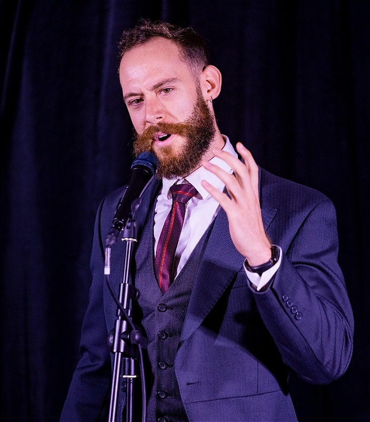 30 y.o bearded man in suit holding his hand out. emotional look on his face. dark room curtains behind, a live production photograph.