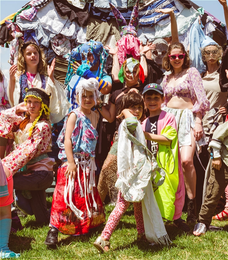 A group of children and adults pose playfully in front of a large sculptural structure made from layers of colourful, draped clothing. Many are dressed in wild, DIY outfits made from fabric scraps, with some wearing masks or headpieces. The mood is energetic and joyful, with bright sun and green grass adding to the festive outdoor setting.