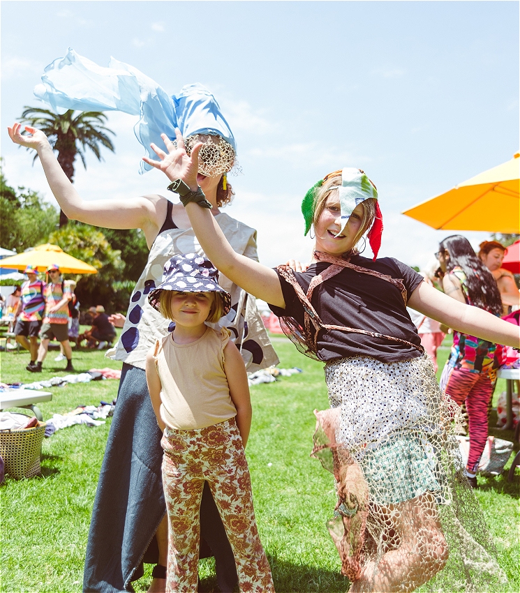 Two children and an adult pose in colourful, makeshift costumes made from fabric scraps and netting on a sunny lawn. One child strikes a dramatic pose, another smiles calmly in floral pants and a bucket hat, while the adult wears a blue veil blowing in the wind. Bright umbrellas and people in costume fill the background.