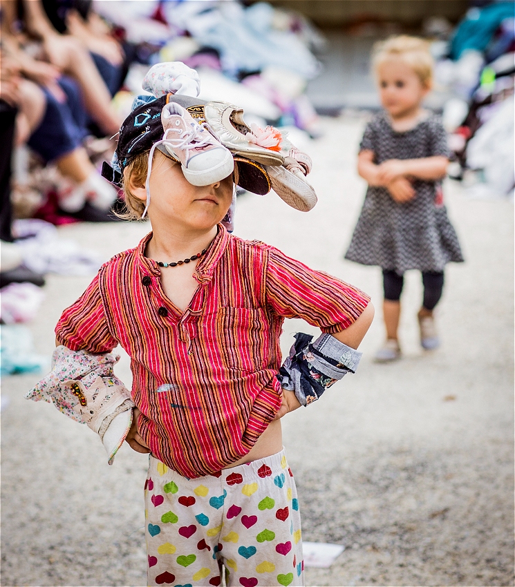 A young child stands confidently on a makeshift runway, hands on hips, wearing a striped red shirt, heart-print pants, and a headpiece made of hats and shoes. Their expression is proud and playful. In the background, another small child watches while adults sit along the runway edges, smiling.