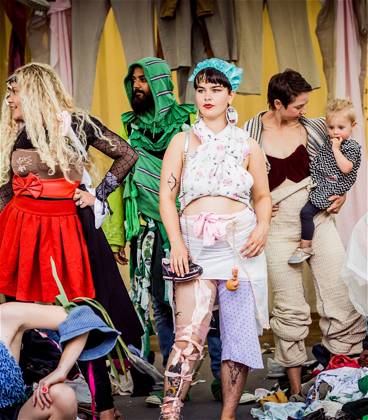 A group of adults and a child pose in a colourful, makeshift fashion scene, wearing outfits created from scraps of fabric, old clothes, and costume pieces. Some strike confident poses, while others smile or interact casually. The backdrop is layered with hanging textiles, and the floor is strewn with clothing waste. The mood is vibrant, playful, and theatrical.