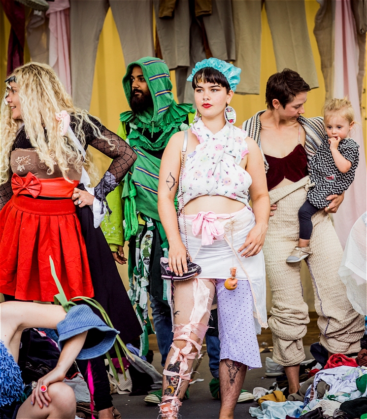 A group of adults and a child pose in a colourful, makeshift fashion scene, wearing outfits created from scraps of fabric, old clothes, and costume pieces. Some strike confident poses, while others smile or interact casually. The backdrop is layered with hanging textiles, and the floor is strewn with clothing waste. The mood is vibrant, playful, and theatrical.