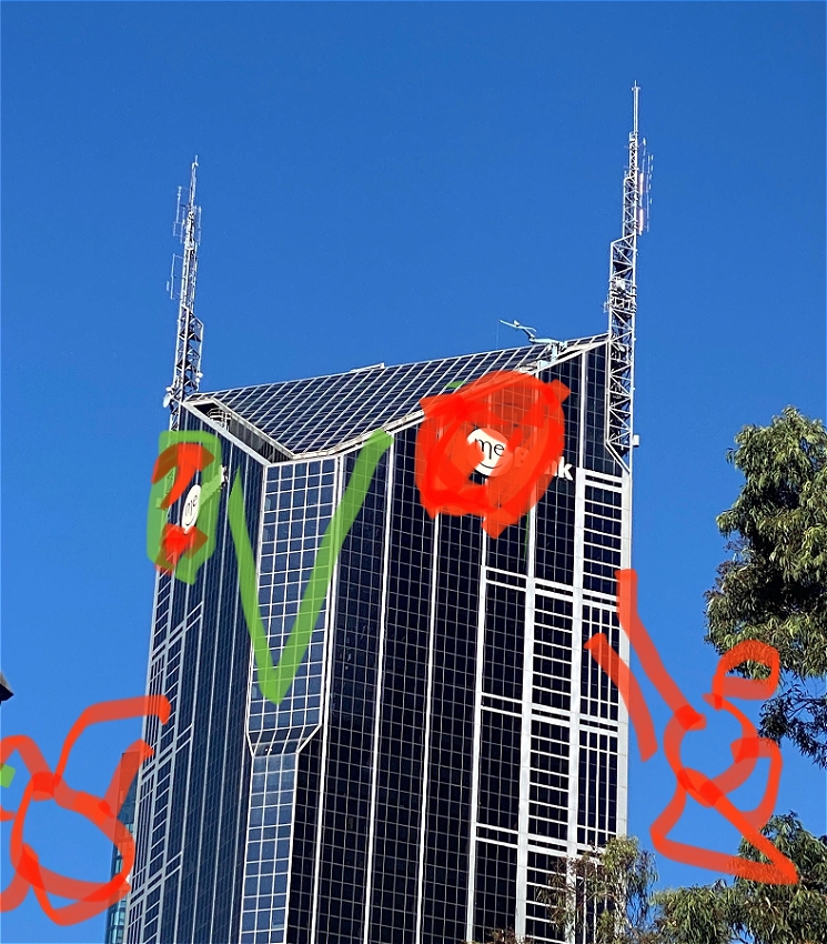 A photograph of black shiny skyscraper seen from below with blue sky behind it. There are child-like scribbled drawings which looks like felt-pen that includes: a green beak, red eyes and red claws. The building has two aerials rising above it that look like horns.