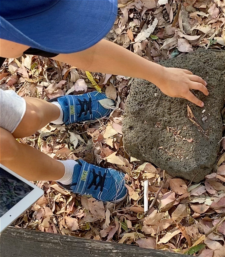 A child seen from above wears blue sneakers and a blue hat. They are holding an iPad and pointing at a rock on the ground.