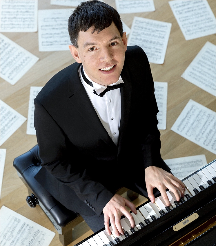 Matthew Shiel is sat at a piano. The image is taken from above with the camera looking down at the subject. The man, Matthew, has brown hair and brown eyes and is playing the piano keys. He is wearing a tuxedo. Behind him you see sheet music thrown across the floor beneath him.